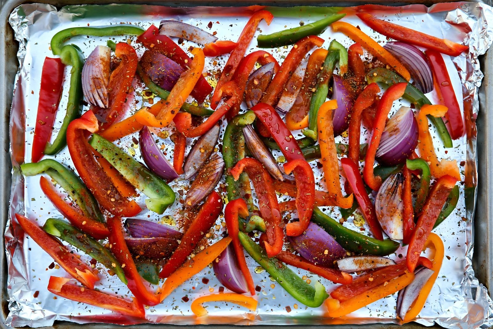 Overhead of seasoned peppers and onion spread in a single layer on a foil-lined sheet pan before roasting.