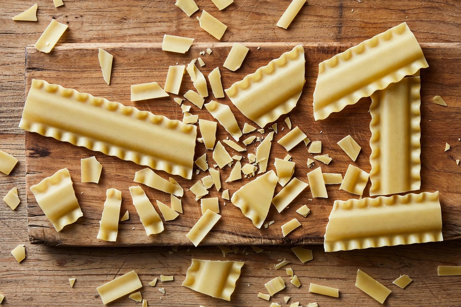 Overhead of lasagna noodles being broken into 1–2 inch pieces over a cutting board.