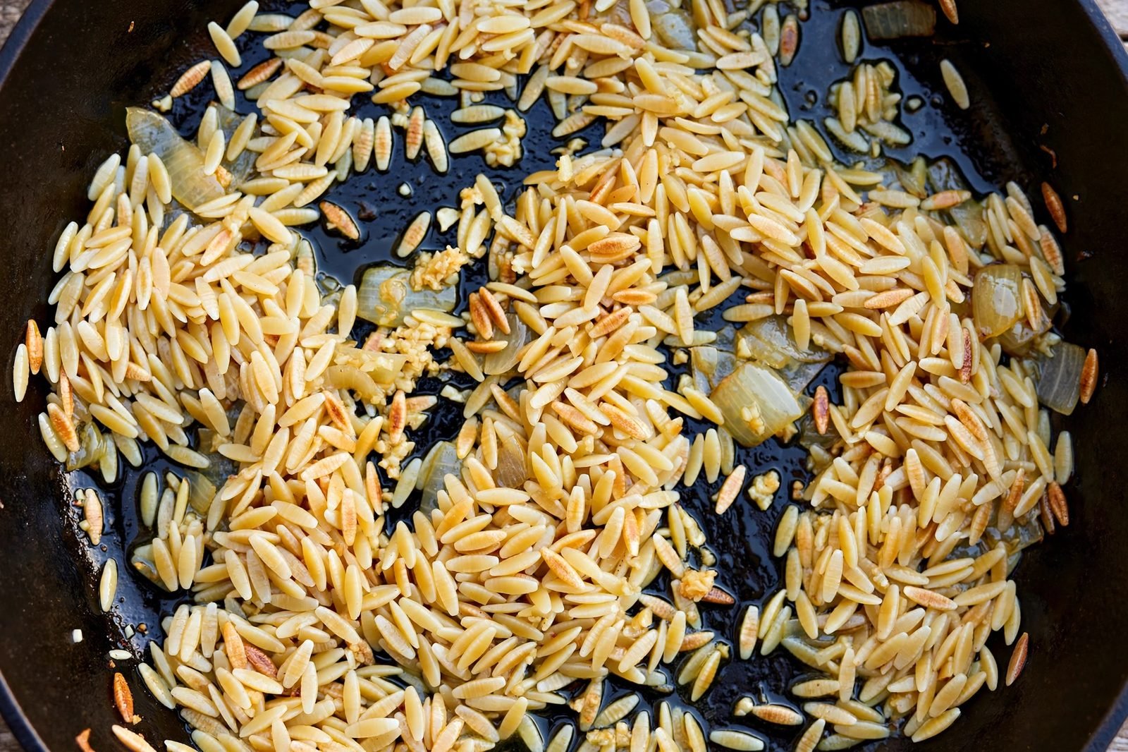 Overhead of dry orzo toasting golden in the skillet with onion and garlic. No people, no text.